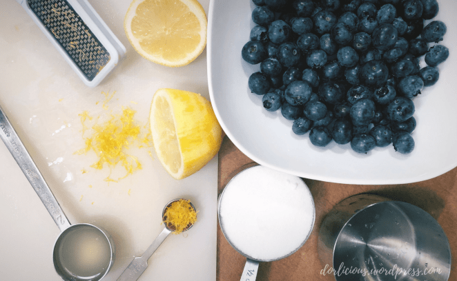 Blueberries in a bowl next to sugar in a measuring cup, a lemon cut in half that has been zested, lemon juice in a measuring spoon, a lemon zester, and lemon zest