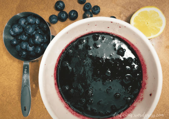 A bowl of blueberry compote next to a measuring cup full of blueberries, spilled blueberries on the counter and half a lemon