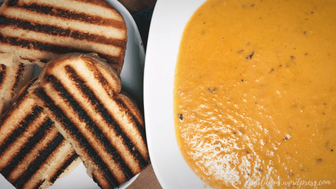 Homemade tomato soup in a white square shaped bowl with rounded corners on a next to grilled cheese with grill marks on it on a white plate