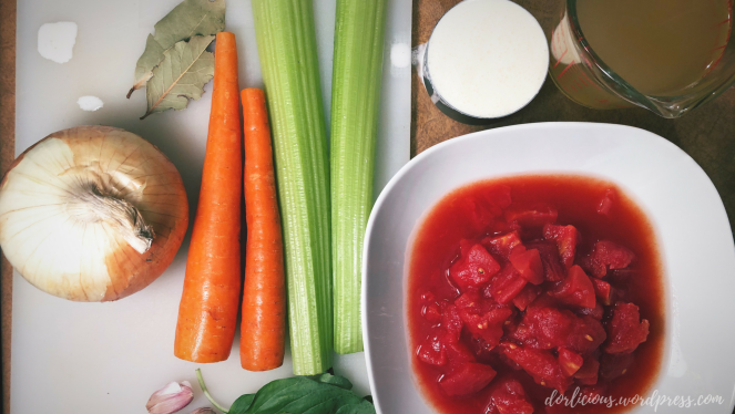 Diced tomatoes in juice, measuring cup of heavy cream, measuring cup of chicken broth, carrots, celery, basil, garlic, an onion and bay leaf on a white cutting board