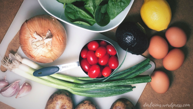 Grape tomatoes, eggs, avocado, lemon, spinach, onion, garlic, scallions and potatoes laying out on a brown counter on top of a white cutting board