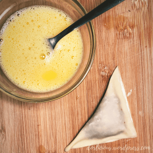 Beaten egg in a small glass bowl with a fork in the bowl next to a prepared potsticker on top of a wooden cutting board