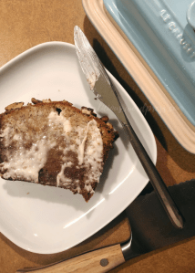 image looking down on a slice of banana bread with butter on a white plate with a knife sitting on the edge of the plate