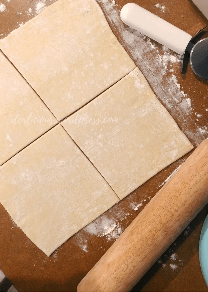 puff pasty that has been rolled out and cut into four squares on a dusted counter next to pizza cutter and rolling pin